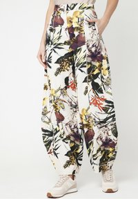 Vrouw in hoge taille, losse bloemmotiefbroek met zijzakken en beige sneakers, staand tegen een effen achtergrond.