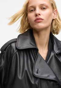 Μαύρο δερμάτινο μπουφάν με διάτρητο σχέδιο, φαρδιά γιακά και κλείσιμο με φερμουάρ. Λείο ύφασμα με εμφανείς ραφές και πολυεπίπεδο σχέδιο.