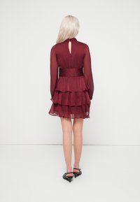 Γυναίκα με μακριά ξανθά μαλλιά, φορώντας ένα στρωτό μπορντό φόρεμα με διαφανείς μακριές μανίκες και μαύρες χαμηλό τακούνι παπούτσια, γυρισμένη προς τα πίσω.