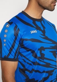 Blaues Polyester-Sportshirt mit schwarzen abstrakten Mustern, kurzen Ärmeln, Rundhalsausschnitt und 'JAKO'-Logo auf der Vorderseite.