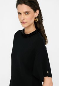 Černý krátký rukáv top s texturovaným černým límečkem, ozdobený zlatým akcentem na rukávu. Modelka nosí zlaté náušnice ve tvaru srdce.