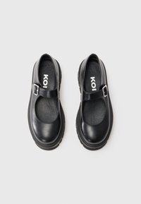 Pair of black Mary Jane shoes with adjustable buckle straps and thick soles on a white background, brand label visible inside.