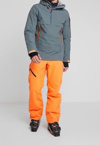 Veste de ski imperméable en teal avec des zips orange, une capuche et une poche poitrine. Associée à un pantalon de neige orange vif, porté avec des bottes de ski noires.