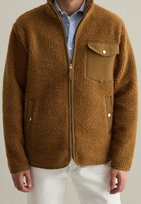 Bruine fleece jas met een ritsluiting, voorzien van een borstzak en zijzakken. Gestructureerde stof met gladde accenten en een relaxte pasvorm.