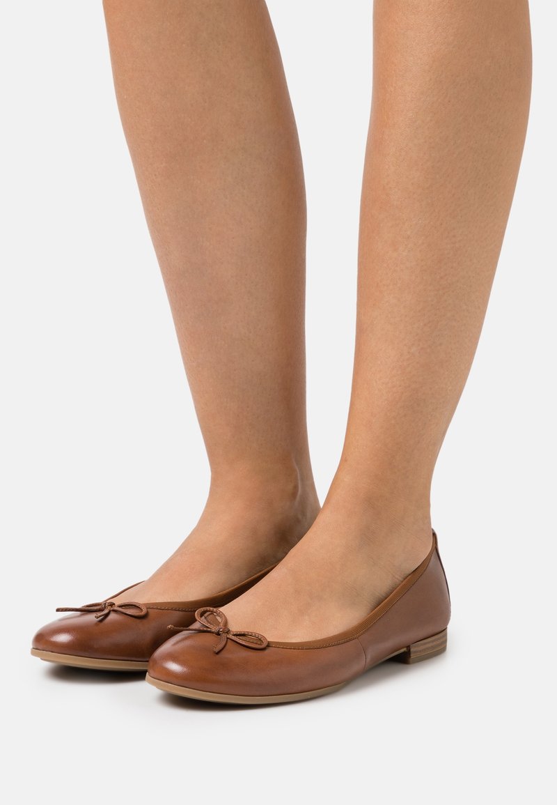 Ballerines en cuir marron avec petits nœuds portées pieds nus sur un fond clair uni.