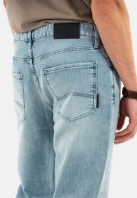 Jeans de mezclilla color azul claro con un acabado ligeramente desvaído, que presenta dos bolsillos traseros y una etiqueta de tela negra en la cinturilla.