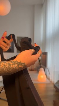 Zwart kledingstuk van stof wordt vastgehouden, met een gladde textuur en vloeiend ontwerp. Lichte achtergrond met een lamp en minimalistische inrichting zichtbaar.