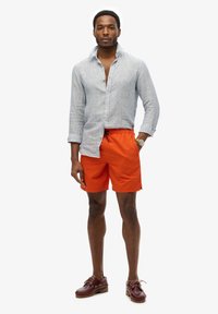 Camisa de lino azul claro con botones, pantalones cortos naranjas con cordón y logotipo grabado, y zapatos de barco burdeos. Hombre de pie con confianza.