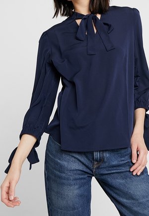 Mujer con blusa azul marino con un lazo atado en el escote y las mangas, combinada con jeans vaqueros azules.