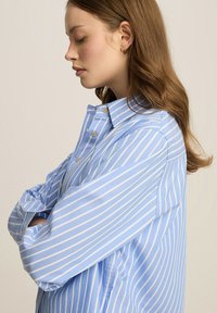 Jasnoniebieska koszula zapinana na guziki z białymi pionowymi paskami, z kołnierzykiem w kształcie spiczastym i długimi rękawami, wykonana z gładkiego, teksturowanego materiału.