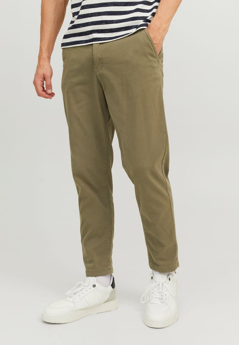 Olivgrüne, schmal zulaufende Hose aus Baumwolle mit Seitentaschen, umgeschlagenen Säumen und kombiniert mit weißen Sneakers und einem gestreiften Hemd.