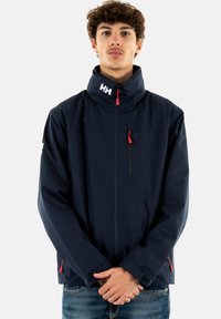 Chaqueta impermeable de color navy con un alto cuello, cremallera frontal y bolsillo en el pecho, con acentos rojos y un logo en el cuello. Textura suave.