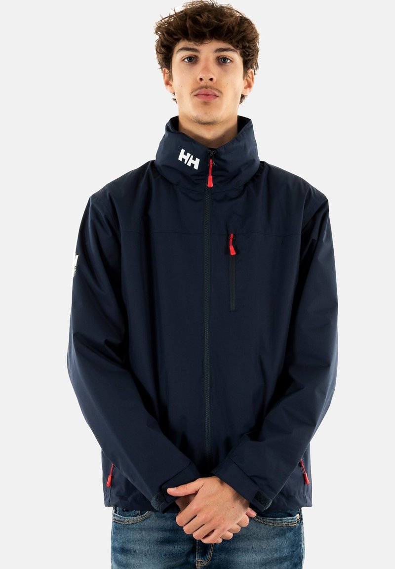 Chaqueta impermeable de color navy con un alto cuello, cremallera frontal y bolsillo en el pecho, con acentos rojos y un logo en el cuello. Textura suave.