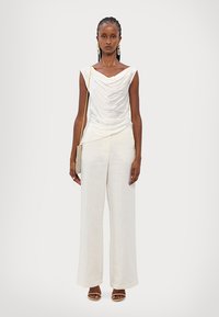 Cowl-neck, sleeveless white top with draped fabric paired with wide-leg, light-colored pants. Model wears strappy sandals and holds a small bag.