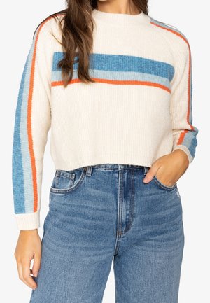 Žena v bílém krátkém svetru s modrými a oranžovými vodorovnými pruhy, v kombinaci s modrými džíny s vysokým pasem, rukou v kapse.