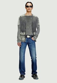 Šedý textúrovaný sveter s náprsníkovými vreckami prehodený cez dlhý rukáv, kombinovaný s modrými džínami a čiernymi teniskami. Doplnky tvoria slnečné okuliare.