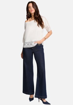 Femme portant un chemisier blanc à épaules dénudées avec dentelle, un jean large et sombre, et des talons pointus foncés, debout devant un fond uni.