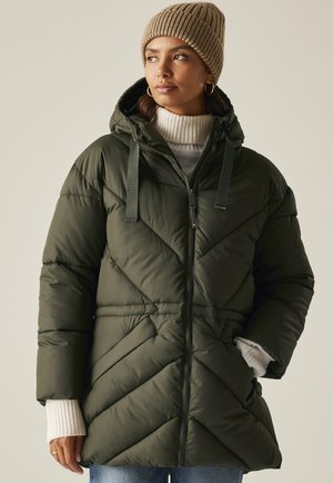 Kobieta ubrana w oliwkowozieloną pikowaną kurtkę z kapturem, beżową dzianinową czapkę, biały sweter z golfem i niebieskie dżinsy, stojąca z rękami w kieszeniach.