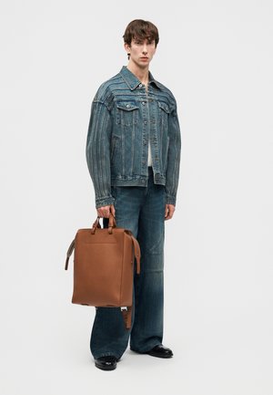 Fiatal férfi túlméretezett kék farmerdzsekit és farmert viselve, egy nagy barna bőr hátizsákot tartva a fogóinál.