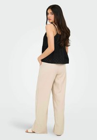 Czarny top bez rękawów o teksturowanym wzorze, w stylu peplum, zestawiony z luźnymi beżowymi spodniami i jasnymi sandałami. Modelka ustawiona bokiem.