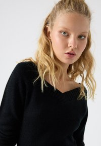 Suéter negro de punto con un amplio escote en forma de V, cuello acanalado texturizado y mangas largas. Tejido suave y ligero con una silueta ajustada.