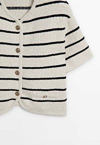 Beżowy sweter w splot z czarnymi paskami, z dekoltem w serek, krótkimi rękawami i teksturowanym materiałem. Posiada okrągłe guziki i małą ozdobną kieszonkę.
