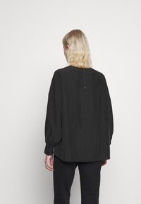 Svart långärmad skjorta med avslappnad passform, med knappar vid nacken baktill och draperade ärmar för extra textur.