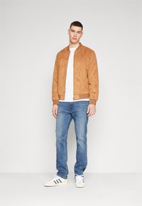Jachetă bomber din suede maro, tricou alb, blugi albaștri și adidași albi cu dungi negre. Modelul stă în picioare într-un cadru neutru.
