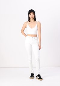 Bijeli crop top i uske bijele traperice u kombinaciji s crnim tenisicama. Osoba nosi crnu kapu i stoji nasuprot jednostavne pozadine.