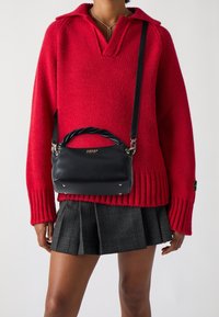 Svart läderhandväska med vridet handtag och silverdetaljer, bärs över en röd stickad tröja och en rutig minikjol.