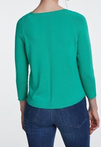 Grüner Strickpullover mit Drei-Viertel-Ärmeln, rundem Ausschnitt und vertikalen Nahtdetails auf der Rückseite. Kombiniert mit dunkelblauen Jeans.