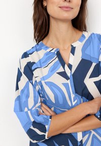 Blusa blu e crema con un motivo a foglia, dotata di scollo rotondo, chiusura con bottoni e maniche 3/4. Realizzata in tessuto leggero con una texture liscia.