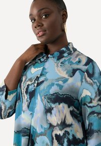Blusa astratta blu con motivo, colletto e chiusura a bottoni. Presenta maniche ampie e una texture di tessuto morbido.