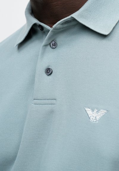 Ανοιχτό μπλε πόλο μπλούζα από υφασμάτινο ύφασμα με γιακά, τρία σκούρα κουμπιά και λευκό κεντημένο λογότυπο στην αριστερή πλευρά.