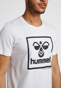 Camiseta de algodón blanca con un gran logo gráfico negro en un cuadrado, con mangas cortas y un escote redondo.