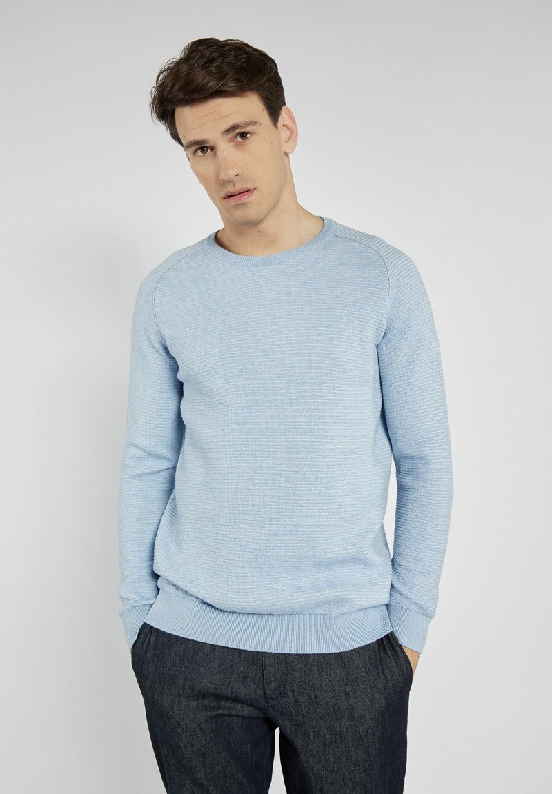 Hellblaue, strukturierte Strickjacke mit langen Ärmeln und Rundhalsausschnitt, kombiniert mit dunkelgrauen Hosen. Einfaches Design, ohne zusätzliche Muster.