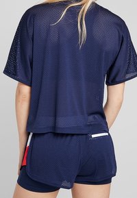 Námořnická modrá sportovní top s volným střihem, krátkými rukávy a kulatým výstřihem, doplněný námořnickými šortkami s červenými a bílými akcenty.