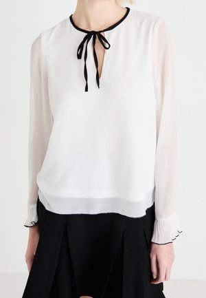 Femme portant une blouse blanche à manches longues avec un bord noir au col et aux poignets, un nœud noir attaché à l'encolure, assortie à une jupe noire.