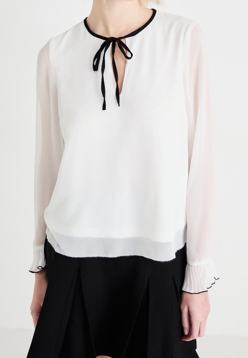 Femme portant une blouse blanche à manches longues avec un bord noir au col et aux poignets, un nœud noir attaché à l'encolure, assortie à une jupe noire.