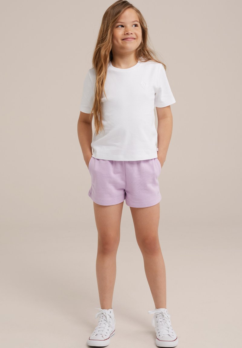 Vit kortärmad T-shirt med ett litet emblem, kombinerad med ljuslila shorts. Modell som bär vita sneakers, med händerna i fickorna.