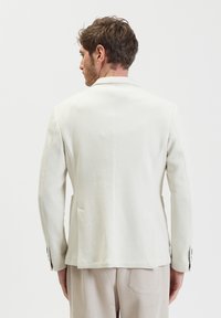 Blazer bianco testurizzato con rever a scocca, tasche laterali e polsini con bottone. Abbinato a pantaloni beige a strisce, visto da dietro.