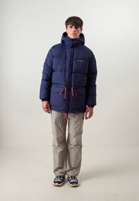 Doudoune marine avec un col haut, deux poches avant et des accents orange vif. Associée à un pantalon gris clair et des chaussures de course.