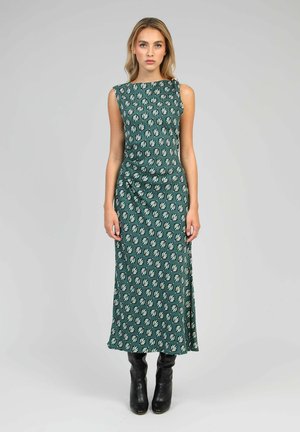 Robe maxi sans manches verte avec un motif de feuilles, fabriquée en tissu doux. Présente un buste ajusté et une taille drapée, portée avec des bottes noires.