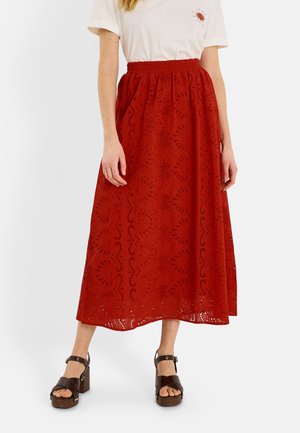 Femme portant une jupe midi rouge à motif œillet avec une ceinture élastique, une chemise blanche à manches courtes et des sandales compensées marron foncé.