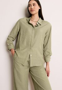 Chemise rayée verte avec col, fermeture à boutons et deux poches avant. Le tissu présente un motif texturé en relief.