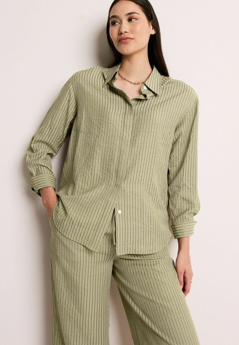Chemise rayée verte avec col, fermeture à boutons et deux poches avant. Le tissu présente un motif texturé en relief.