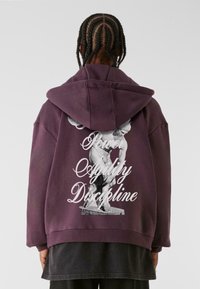 Sudadera morada de gran tamaño con capucha ajustable, que presenta un gráfico en blanco y negro de una estatua y el texto: "Poder Agilidad Disciplina" en la parte posterior.