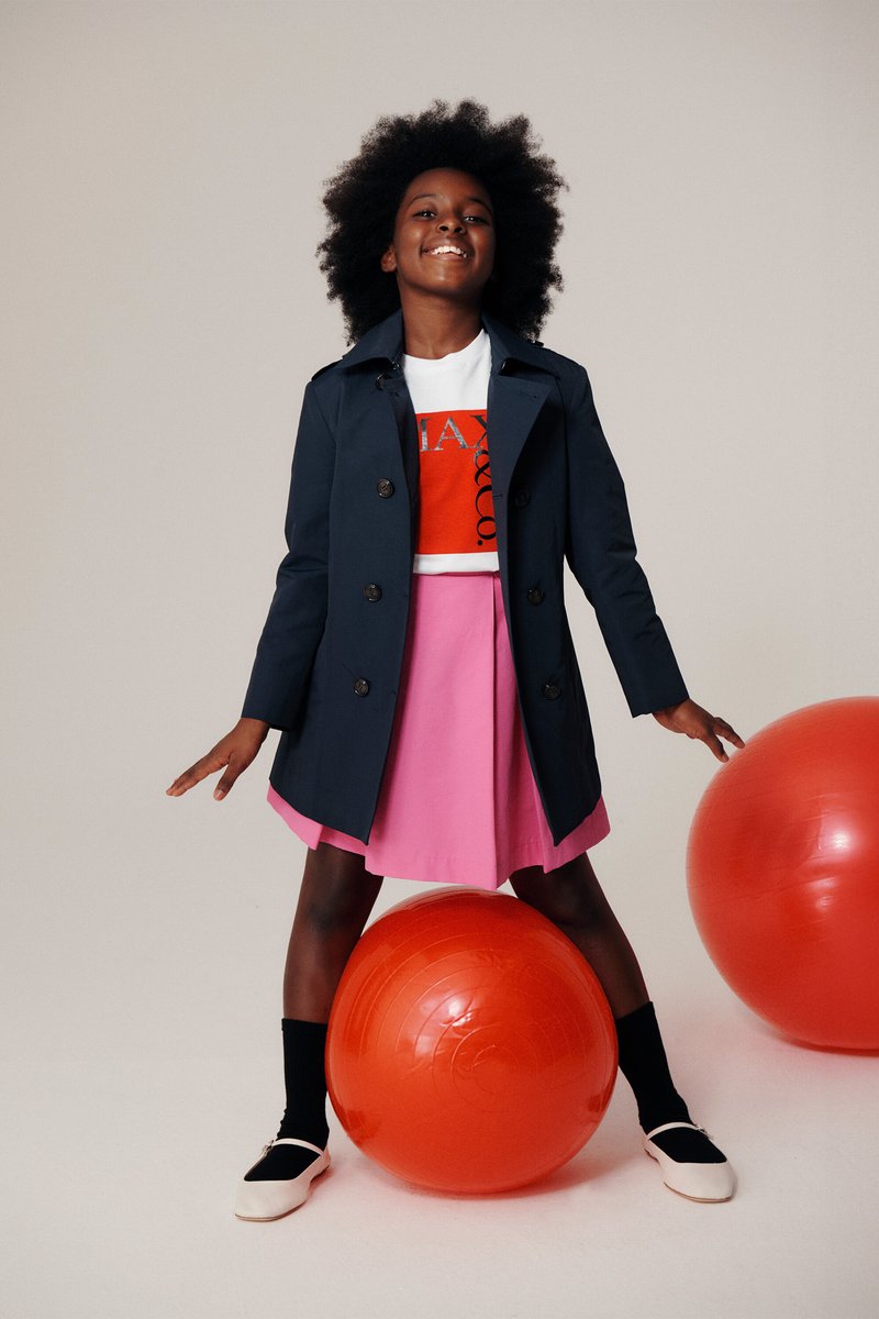 Manteau trench marine, chemise blanche avec un graphique rouge, jupe rose, chaussettes noires jusqu'aux genoux et chaussures noires et blanches. Pose avec des ballons de gym rouges.