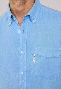Camicia azzurra chiara con tessuto testurizzato, colletto a punta e una tasca sul petto con un'etichetta. Quattro bottoni bianchi sul davanti.