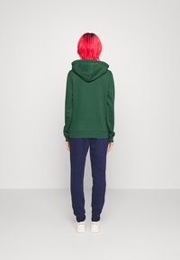 Sudadera verde con capucha y bolsillos laterales, combinada con pantalones jogger azul marino. El modelo lleva zapatillas blancas y se ve desde atrás.
